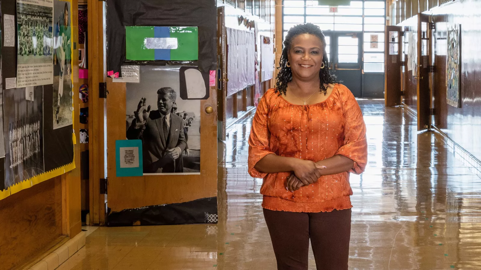 A teacher in an orange shirt stands smiling in a very bright and clean high school hallway.