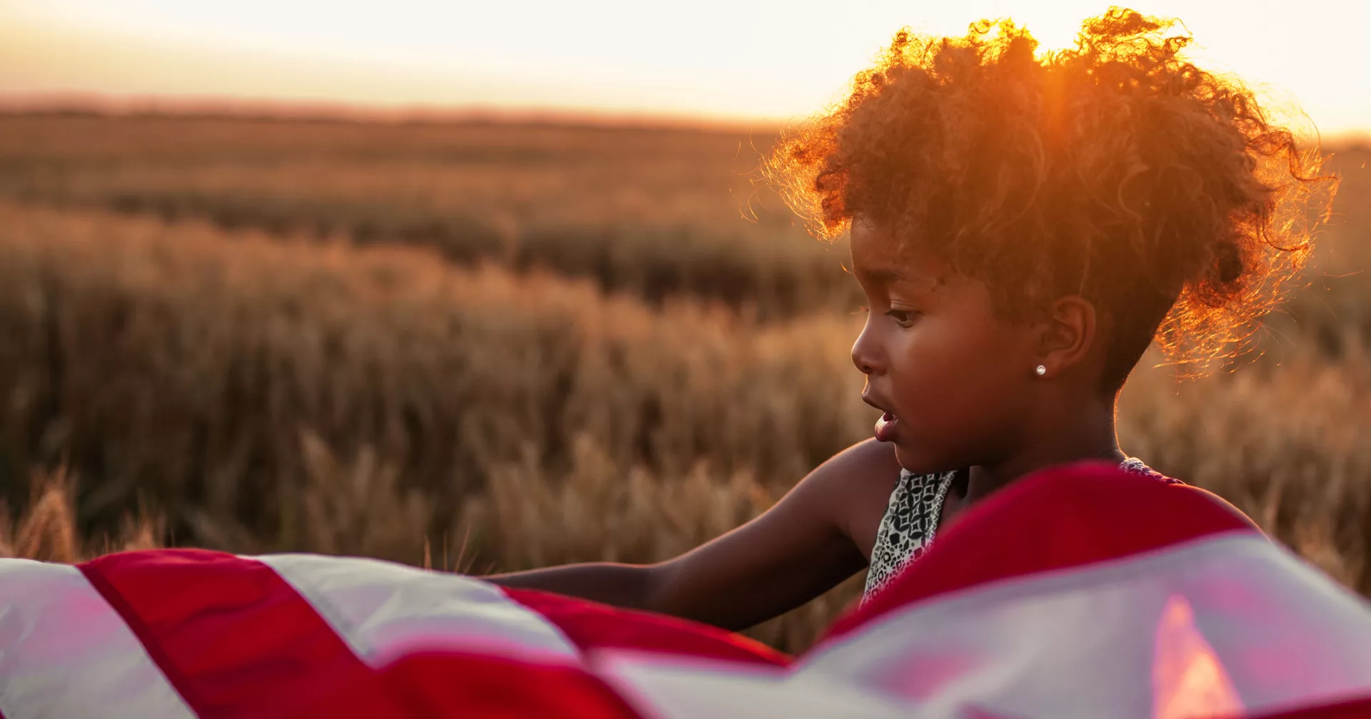 the sun sets behind a young Black girl in a field with an American flag blowing in the wind